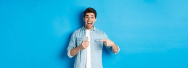 Çevrimiçi alışveriş, uygulama ve teknoloji kavramı. Mutlu gülümseyen adam telefonu işaret ediyor, mavi arka plana hayret içinde bakıyor..