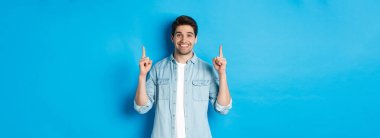 Sıradan kıyafetli yakışıklı sakallı bir adamın portresi, gülümseyen mutlu ve parmaklarını fotokopi alanına gösteren, mavi arka planda duran.