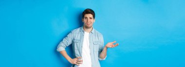 Dikkatsiz bir adam hayal kırıklığı yaratan bir şeye bakıyor, şikayet ediyor ve kötü ürünü yargılıyor, mavi arka planda eğlenmeden duruyor..