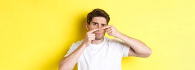 Genç adam burnuna sivilce yapıyor, sarı arka planda duruyor. Cilt bakımı ve sivilce kavramı.