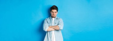 Öfkeli adam göğsünde çapraz silah taşıyor ve aşağılanmış bir ifadeyle yüzünü çeviriyor. Mavi arka plana karşı kırgın duruyor..