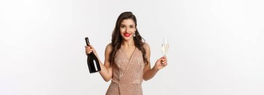 Kış bayramı kutlaması. Lüks elbiseli zarif bir kadın, yeni yıl partisinde içiyor, elinde bardak ve bir şişe şampanya, beyaz arka planda duruyor..