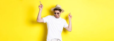 Turizm, seyahat ve tatil konsepti. Erkek turist tatilden zevk alıyor, hasır şapka ve güneş gözlükleriyle dans ediyor, parmaklarını yana doğru gösteriyor..