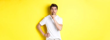 Heyecanlı bir adamın sürpriz planlaması, kameraya şşşş yapması, sessiz kalmak için parmağa basması, sarı arka planda durması..