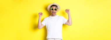 Turizm ve yaşam tarzı kavramı. Mutlu adam tatil köyüne gidiyor, evet diye bağırıyor ve ellerini kaldırıyor, zafer kazanıyor, güneş gözlüğü takıyor ve yaz şapkası takıyor, sarı arka plan.