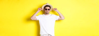 Tatile hazır, hasır şapka ve güneş gözlüğü takmış, sarı arka planda duran kendinden emin genç turist..