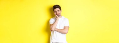 Sıkılmış ve keyifsiz bir adam, kayıtsız bakıyor, sarı arka planda beyaz elbiseler içinde duruyor..