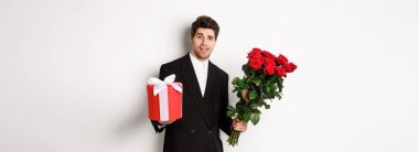 Tatil, ilişki ve kutlama kavramı. Siyah takım elbiseli yakışıklı ve kendinden emin bir adam, randevuya çıkıyor, elinde gül buketi, beyaz arka planda duruyor..