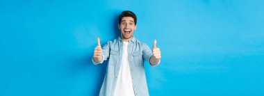 Gülümseyen kendine güvenen bir adam heyecanlı bir yüz ifadesiyle, harika bir şey gibi, ürünü onaylayan, mavi arka plana karşı duran.