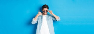 Güneş gözlüklü yakışıklı maço adam seninle flört ediyor, telefon işareti yapıyor ve göz kırpıyor, bana jest diyor, mavi arka planda duruyor..