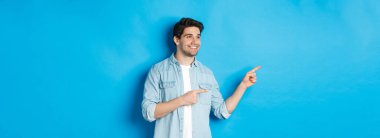 Yakışıklı yetişkin adam ürünü tanıtır, bakar ve işaret eder, mavi arka plana karşı bir şeyi teşvik eder..