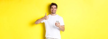 Akıllı telefon tutan mutlu bir adam, onaylanmış bir şekilde baş parmağını gösteriyor, sarı arka planda duran, çevrimiçi bir şey öneriyor..