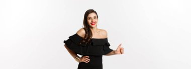 Moda ve güzellik. Cazip elbiseli, kırmızı rujlu genç ve çekici bir kadın. Başparmağını göstererek onaylıyor. Bir şey öneriyor..