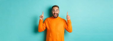 Young happy man showing advertisement, pointing fingers up announcing promo offer, standing over turquoise background.