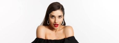 Zarif küpeler ve siyah elbiseler giyen muhteşem esmer kadının yakın plan çekimi. Gözünü delip geçen bakışlarla kameraya şehvetli bir şekilde bakıyor ve beyaz arka planda duran ağzı açık bir kadın..