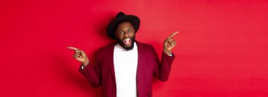 Kış tatili ve alışveriş konsepti. Neşeli siyahi adam gülümsüyor ve iki reklam gösteriyor, fotokopi alanlarını işaret ediyor, kırmızı arka plan.