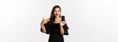 İnternetten alışveriş konsepti. Siyah elbiseli şık bir kadın akıllı telefon ekranını işaret ediyor. Uygulamayı gösteriyor. Beyaz arka planda duruyor..