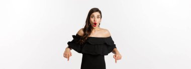 Moda ve güzellik. Siyah elbiseli zarif bir kadın, parmakları aşağıda, tanıtım yapıyor ve gülümsüyor, beyaz arka planda duruyor..