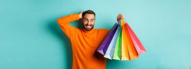 Yakışıklı genç adam garip ve kaşınan kafa, hediye dolu alışveriş çantalarına bakıyor, turkuaz arka planda dikiliyor..