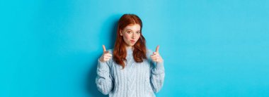 Gururlu kızıl kız kamerayı işaret ediyor, övgüler yağdırıyor ve tebrik ediyor, mavi arka planda duruyor. Boşluğu kopyala