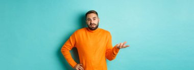 Dikkatsiz ve umursamaz adam omuz silkiyor, rahatsız edilmemiş görünüyor, hiçbir şey bilmiyor, açık mavi arka planda duruyor..