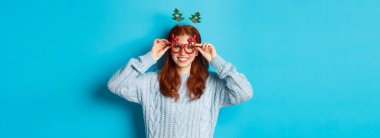 Kış tatili ve Noel satış konsepti. Güzel kızıl saçlı manken, yeni yılı kutluyor, komik bir saç bandı ve gözlük takıyor, kameraya gülümsüyor..