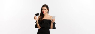 Moda ve alışveriş konsepti. Kırmızı dudaklı çekici bir kadın, siyah elbiseli, ne alacağını düşünen, kredi kartı ve cep telefonu tutan, beyaz arka planda duran.