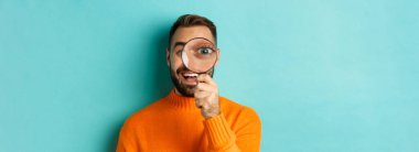 Komik adam büyüteçten bakıyor, bir şeyi arıyor ya da araştırıyor turkuaz arka planda turuncu süveter giyiyor..