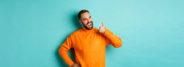 Mutlu erkek model baş parmağını gösteriyor, sol üst köşedeki logodan memnun görünüyor, ürünü onaylıyor ve tavsiye ediyor, açık mavi arka planda duruyor..