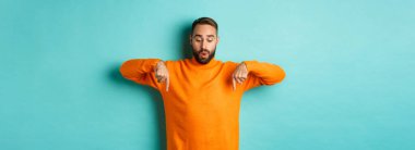 Turuncu süveter giyen, parmaklarını aşağıya gösteren, reklamlara ilgi duyan, tatil indirimleri yapan, turkuaz arka planda duran harika adam..