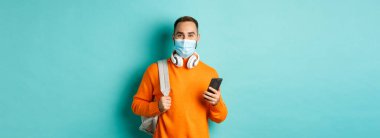 Yüz maskeli genç adam cep telefonu kullanıyor, sırt çantası taşıyor, açık mavi arka planda duruyor. Boşluğu kopyala