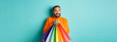 Turuncu kazaklı mutlu beyaz adam, sola bakıyor ve görüntülüyor, alışveriş torbaları tutuyor ve gülümsüyor, turkuaz arka planda dikiliyor..