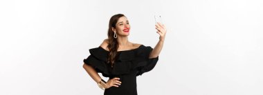 Siyah elbiseli güzel bir kadın partide selfie çekiyor, beyaz arka planda dikiliyor, akıllı telefonlu..