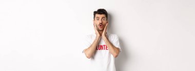 Gönüllü tişörtlü şok olmuş bir adam, hayranlıkla kameraya bakıyor, beyaz arka planda duruyor..