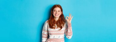 Sevimli, gülümseyen kızıl saçlı kadın, iyi bir işaret veriyor, iyi iş çıkartıyor, iyi bir jest yapıyor, mavi arka planda duruyor..