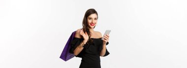 Güzellik ve alışveriş konsepti. Zarif siyah elbiseli ve makyajlı güzel bir kadın, cep telefonundan sipariş veriyor, çantasıyla gülümsüyor, beyaz arka planda dikiliyor..