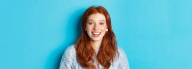 Kazaklı mutlu kızın yakın çekimi, umut dolu bir gülümsemeyle kameraya bakıyor, mavi arka planda duruyor..