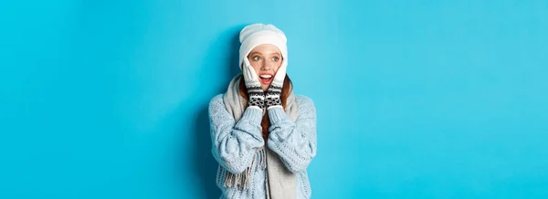 Excited redhead girl staring left at logo, wearing winter clothes, beanie, gloves and sweater, standing over blue background.