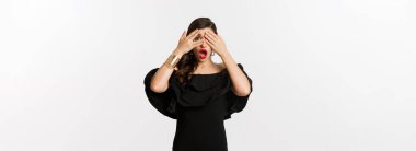 Moda ve güzellik. Şok olmuş, siyah elbiseli, gözleri kapayan ama parmaklarından bakan beyaz arka planda duran büyüleyici kadının görüntüsü..