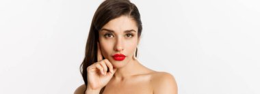 Moda ve güzellik konsepti. Siyah elbiseli, gece makyajlı ve kırmızı rujlu zarif güzel bir kadın, kameraya küstah bakıyor, beyaz arka planda duruyor..