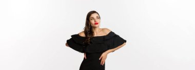 Moda ve güzellik. Siyah elbiseli, makyajlı, kırmızı dudaklı, beyaz arka planda kendine güvenen, beyaz arka planda duran modern bir kadın..