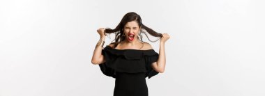 Moda ve güzellik. Siyah elbiseli genç bir kadın çığlık atıyor, saçlarını başını yoluyor, deli gibi bağırıyor, kızgın ve öfkeli bir şekilde beyaz arka planda duruyor..