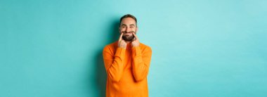 Sakallı bir adamın mutlu bir gülümsemeyle dudaklarını germesi, sahte mutluluk, açık mavi arka planda durması. Boşluğu kopyala