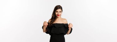 Moda ve güzellik. Siyah elbiseli zarif bir kadın, parmakları aşağıda, tanıtım yapıyor ve gülümsüyor, beyaz arka planda duruyor..