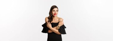 Moda ve güzellik. Siyah elbiseli güzel, çekici bir kadın, seçim yapıyor, baştan çıkarıcı dudağını ısırıyor ve yan tarafları işaret ediyor, beyaz arka planda duruyor..