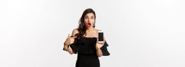 İnternetten alışveriş konsepti. Siyah elbiseli şık bir kadın akıllı telefon ekranını işaret ediyor. Uygulamayı gösteriyor. Beyaz arka planda duruyor..
