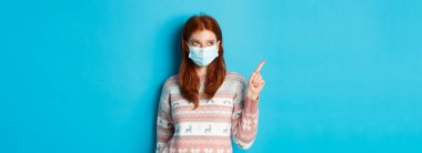 Kış, covid-19 ve karantina konsepti. Yüz maskesi ve süveter takmış, sol üst köşeyi gösteren, ürünü seçen, mavi arka planda duran kızıl saçlı güzel bir kız resmi..