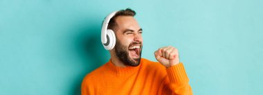 Kulaklıkla müzik dinleyen, turuncu süveter giymiş turkuaz arka planda duran yakışıklı modern adamın yakın çekimi..