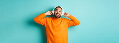 Turuncu süveter giyen mutlu yetişkin adam, yukarı bakıyor ve kulaklıkla müzik dinliyor, mavi arka planda duruyor..