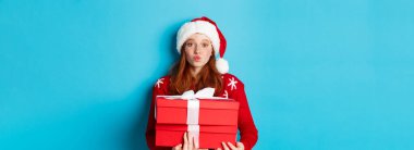 Mutlu tatiller ve Noel konsepti. Sevimli kızıl kız hediyeler tutuyor ve öpücük için dudaklarını büzüştürüyor, Noel Baba şapkası ve komik süveter giyiyor, mavi arka plan.